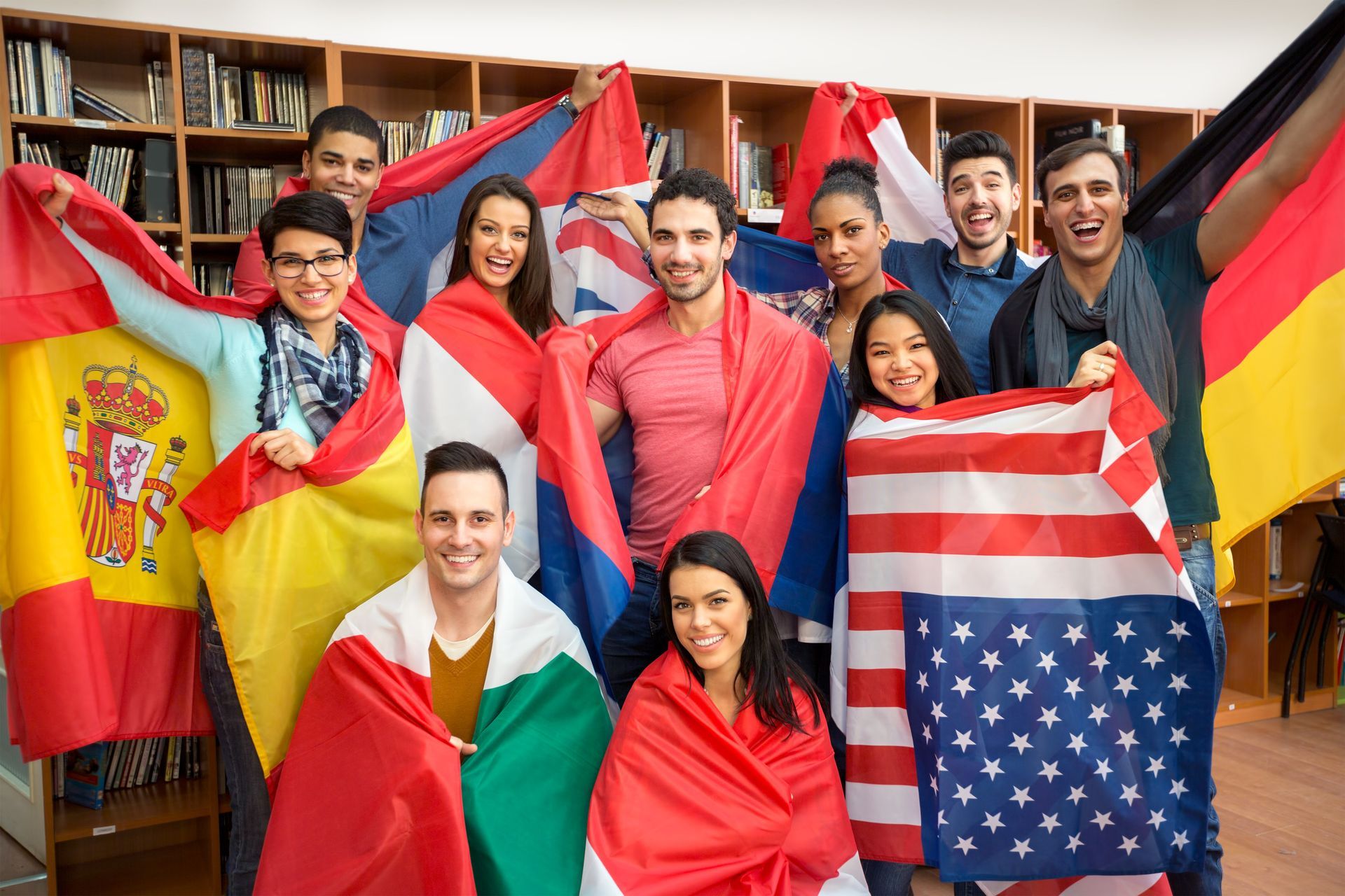 Grupo diverso de estudiantes envueltos en banderas, sonriendo en una biblioteca.