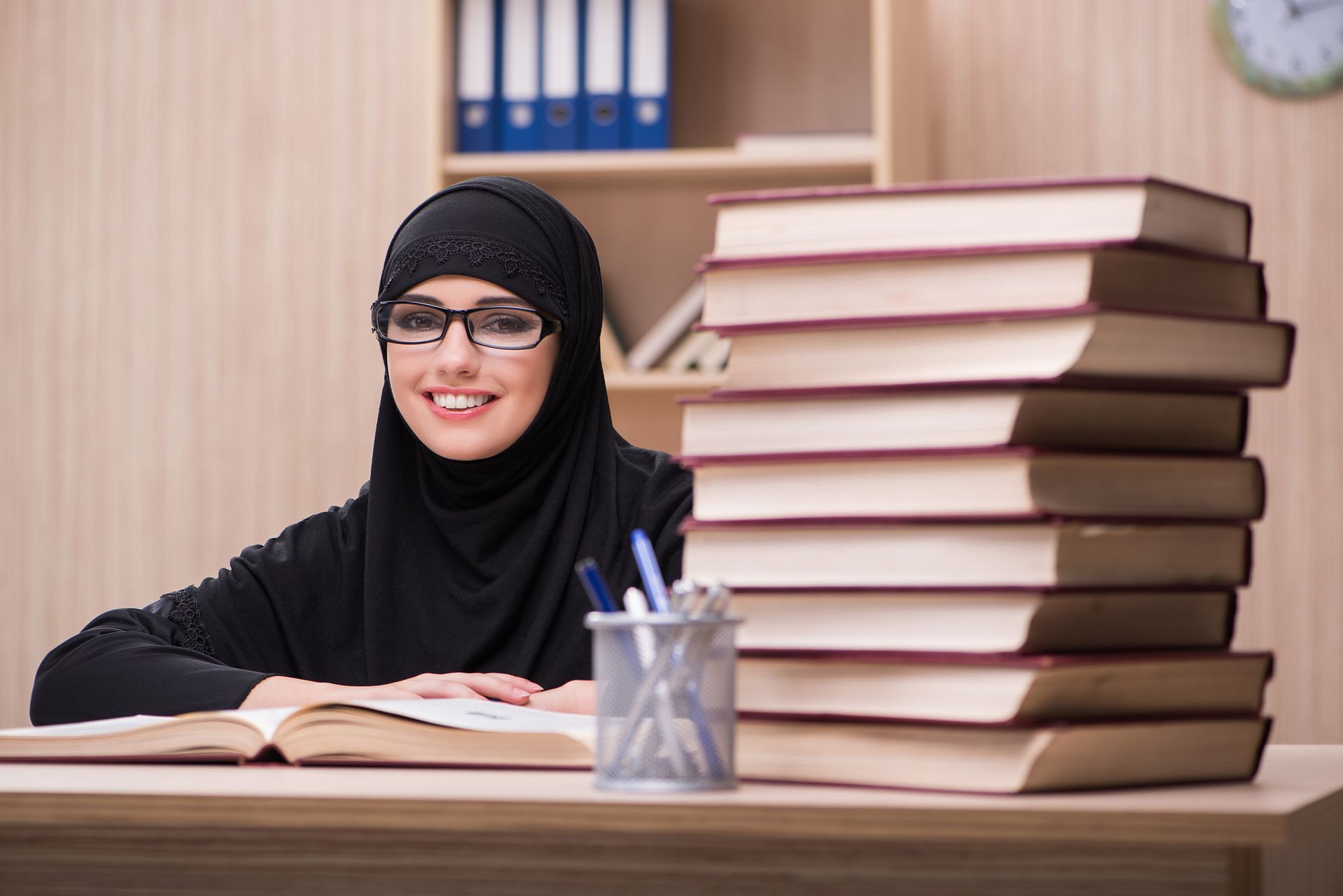 Una mujer con hijab y gafas sonríe, leyendo en un escritorio, con una pila de libros y un portalápices.