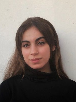 Una mujer de pelo largo y castaño, vestida con un cuello alto negro, sonríe a la cámara frente a una pared blanca.