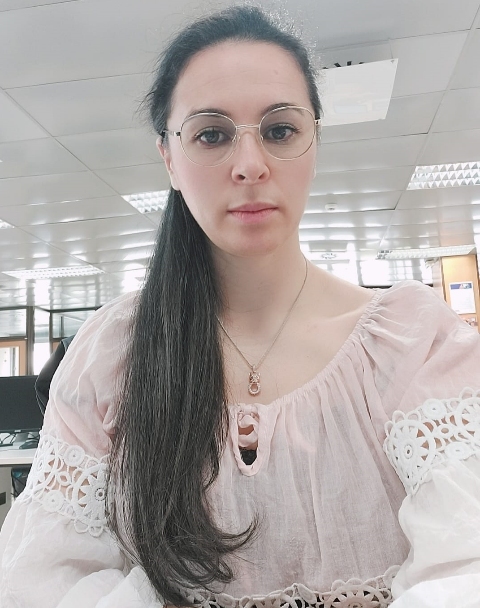 Mujer con gafas y cola de caballo larga con blusa rosa, en el interior, mirando a la cámara.