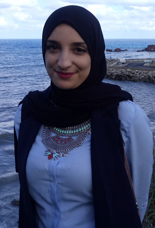 Mujer con hijab negro y chaleco, sonriendo, al aire libre junto al océano, luciendo un collar.