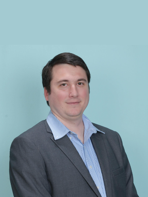 A person with short brown hair wearing a light blue striped dress shirt and a grey blazer against a light blue background.