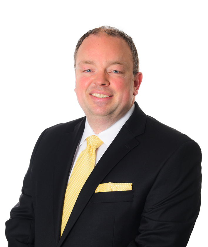 Headshot of a smiling person wearing a dark suit, white shirt, and a yellow patterned tie with a matching pocket square.