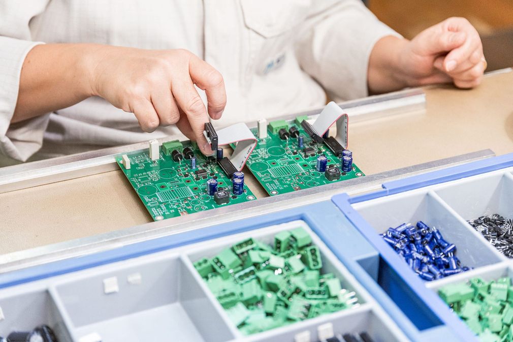 Des mains assemblent des cartes de circuits imprimés vertes au-dessus de bacs de composants électroniques sur un établi.