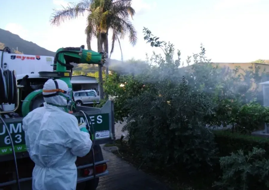 Persona con traje de protección rociando un sistema de pesticida montado en un vehículo sobre arbustos