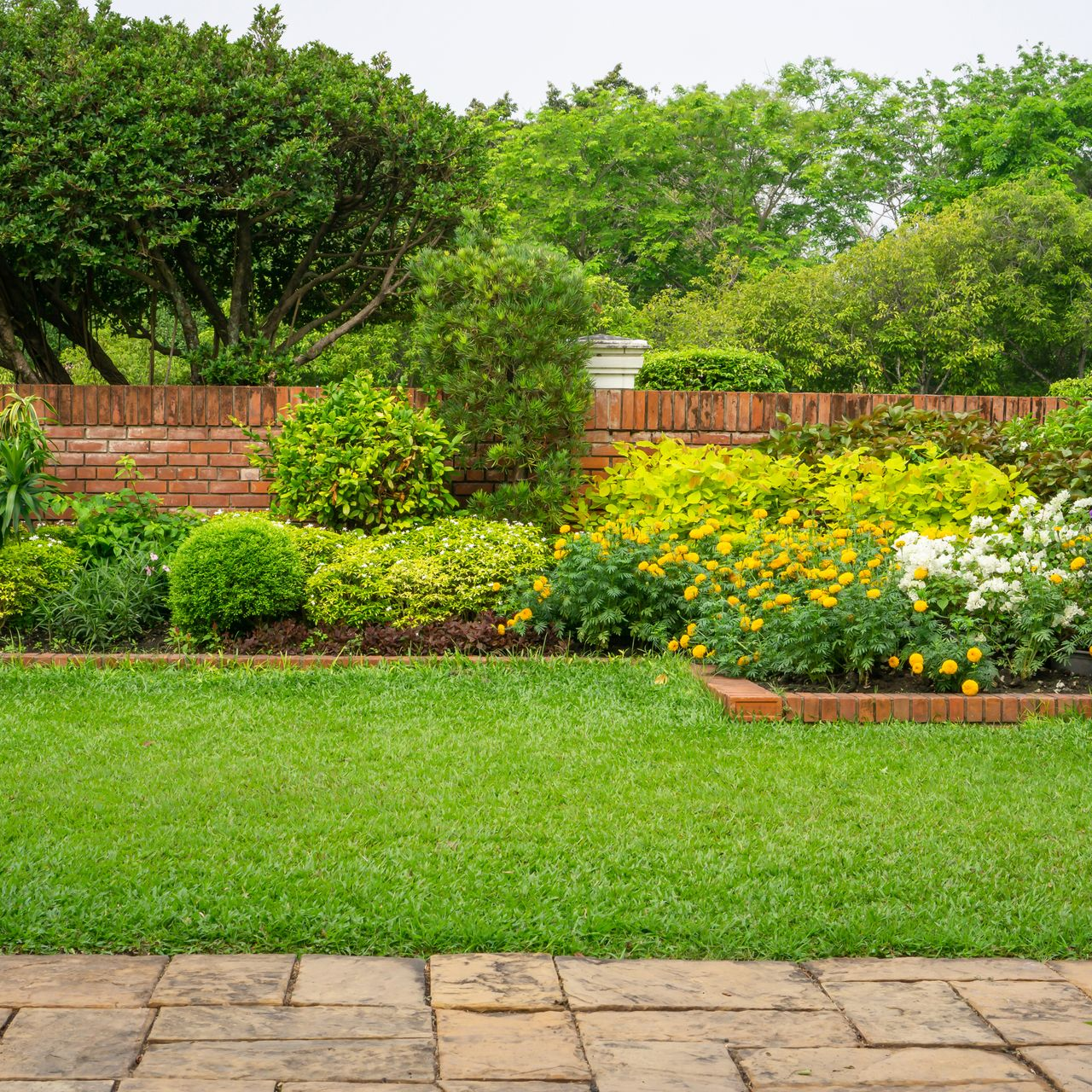 Jardin luxuriant avec une pelouse verte vibrante, des bordures en briques et des fleurs colorées en fleurs contre un mur de briques.