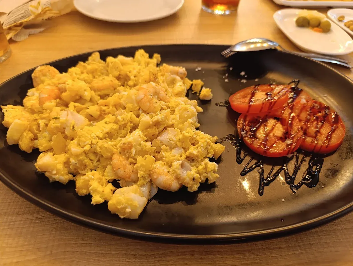 Huevos revueltos con camarones y tomates a la parrilla en un plato negro, rociados con glaseado balsámico.