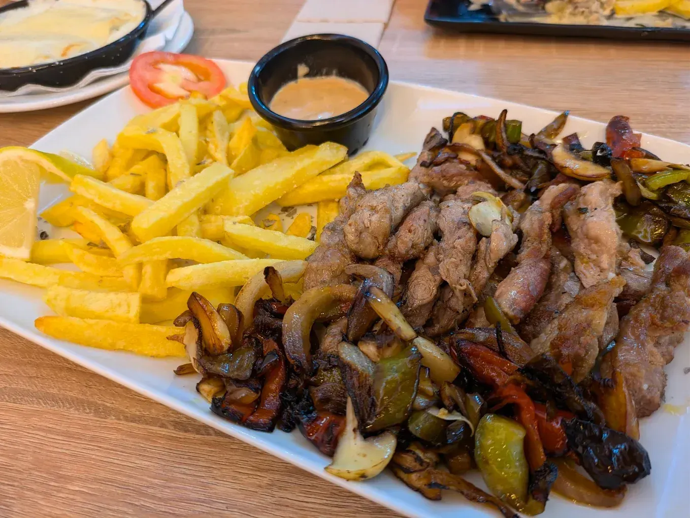 Plato de comida: carne en rodajas con pimientos salteados, patatas fritas, salsa y limón.