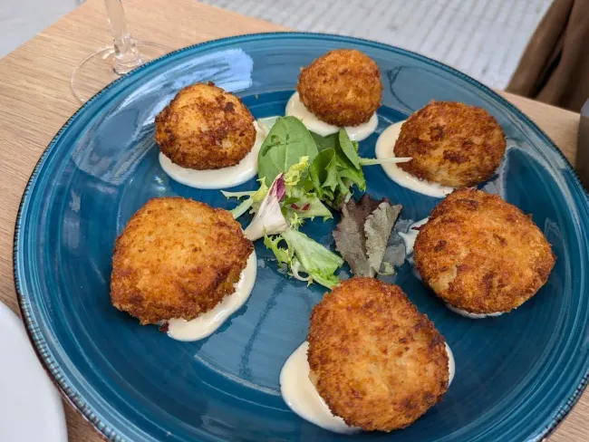Seis croquetas doradas y fritas en un plato azul, cada una sobre una cucharada de salsa blanca y adornadas con verduras.