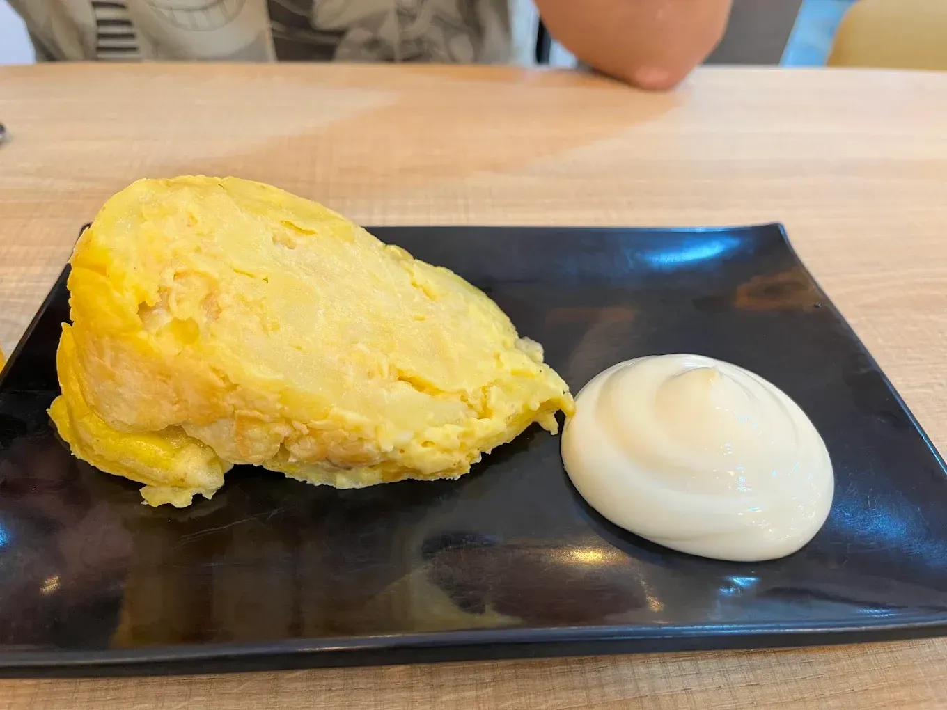 Tortilla de patatas con salsa blanca en un plato negro, sobre una mesa de madera.