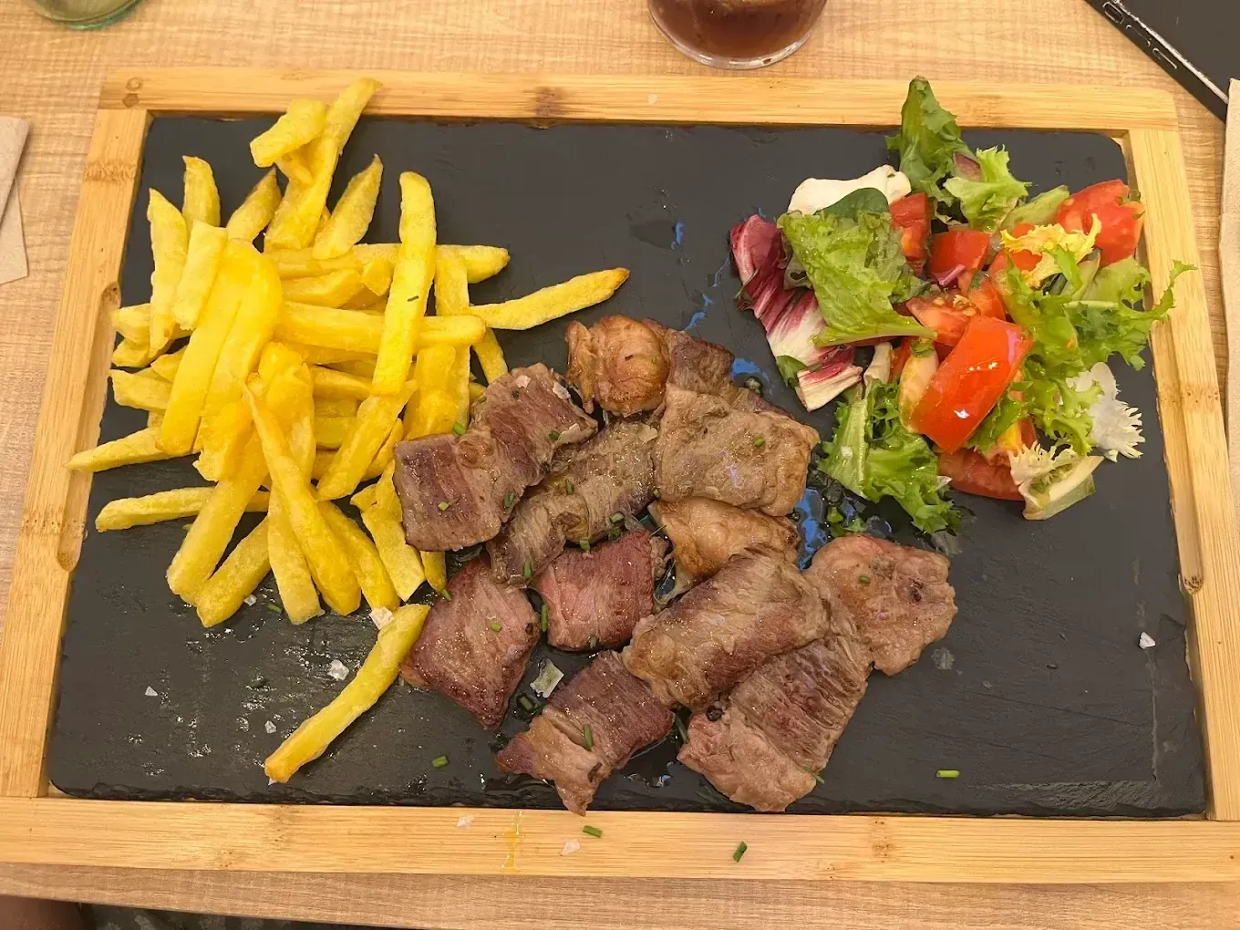 Cubos de carne, papas fritas y ensalada sobre una tabla para servir de pizarra negra con borde de madera.