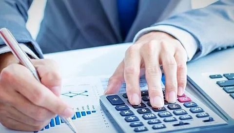 Persona de traje usando calculadora y escribiendo en un documento financiero con un bolígrafo.