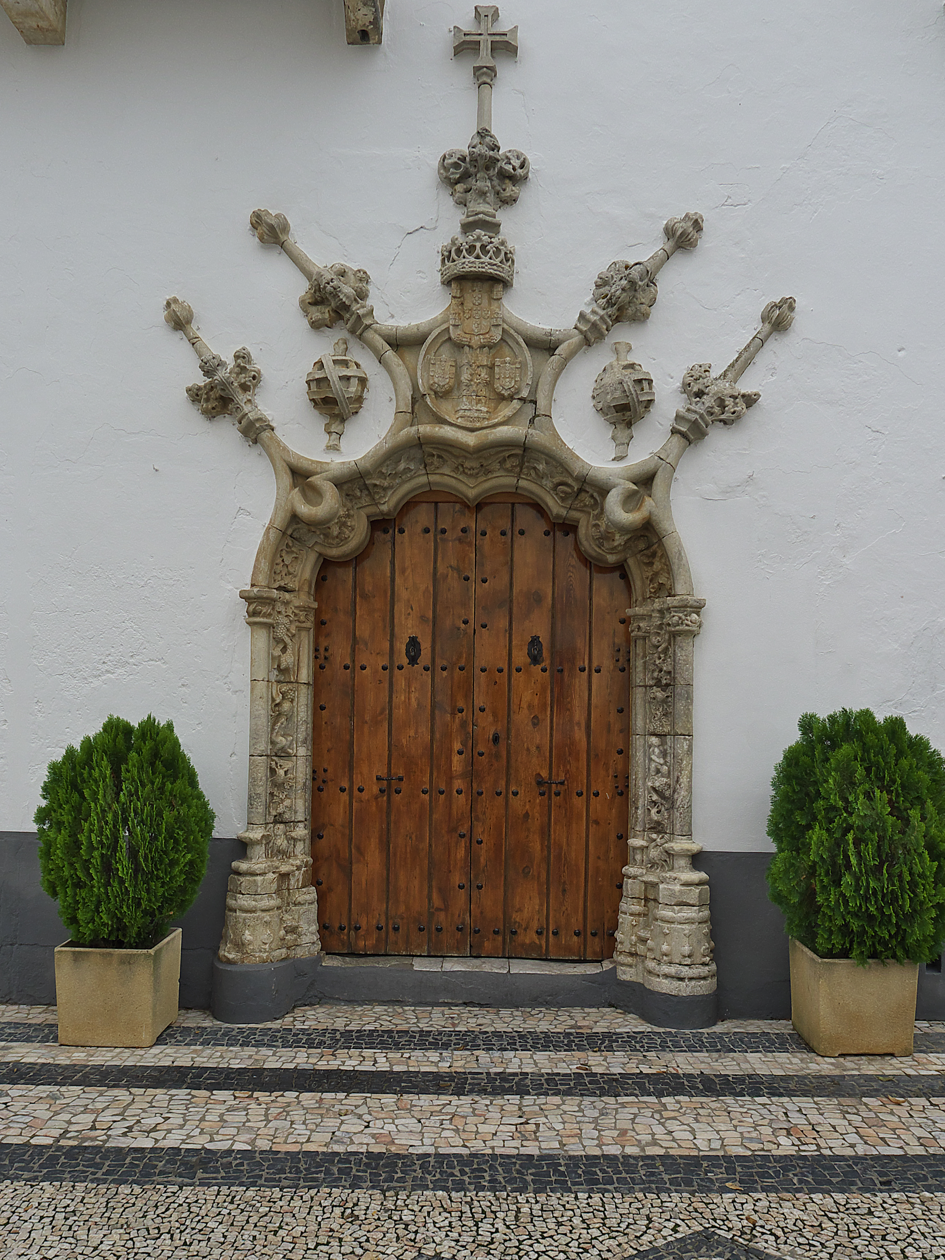 Puerta de iglesia de madera adornada con tallas de piedra, cruz y plantas en macetas a los lados.