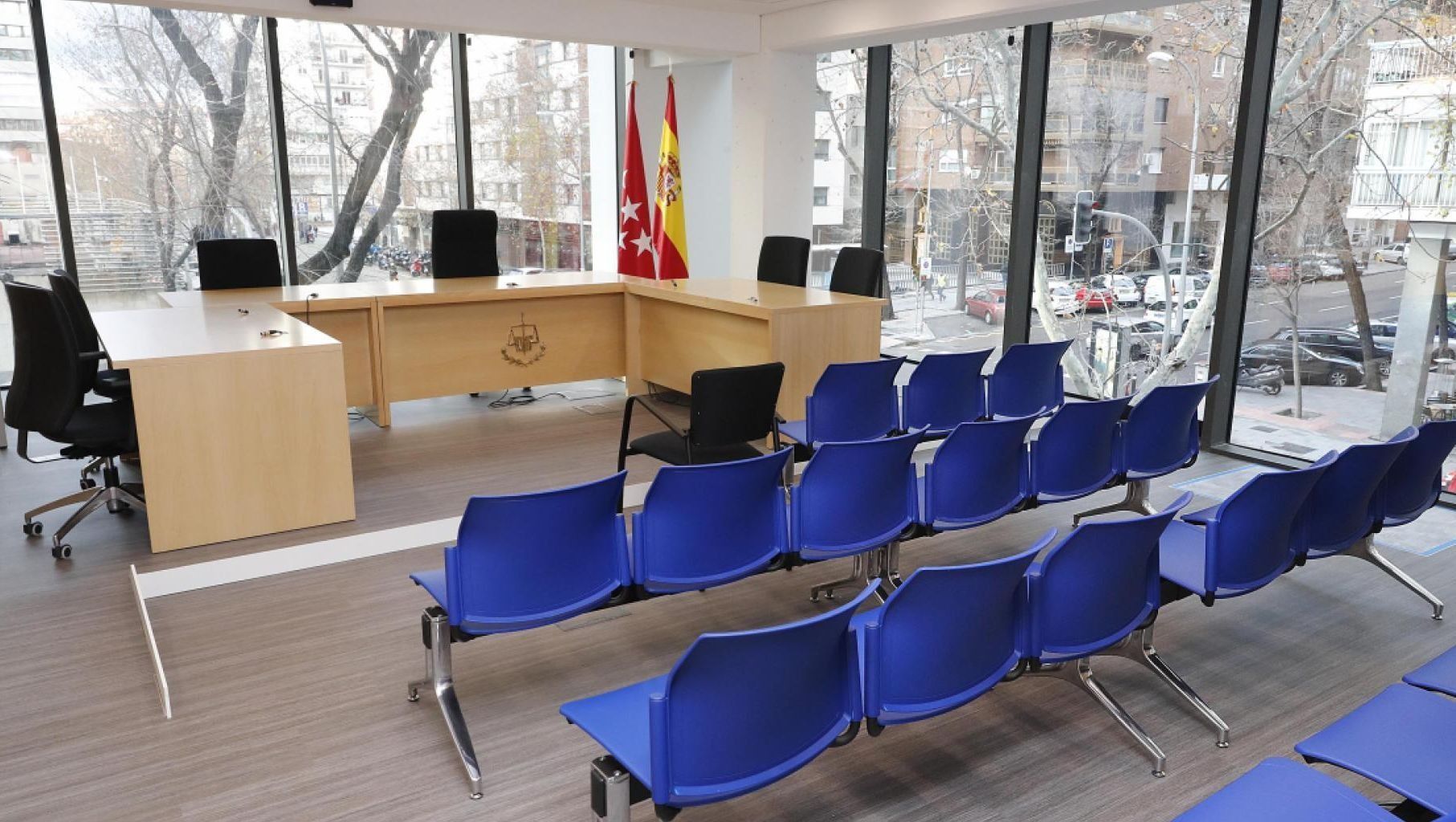 Una sala de audiencias moderna con filas de sillas azules frente a un escritorio de madera, una ventana y dos banderas.
