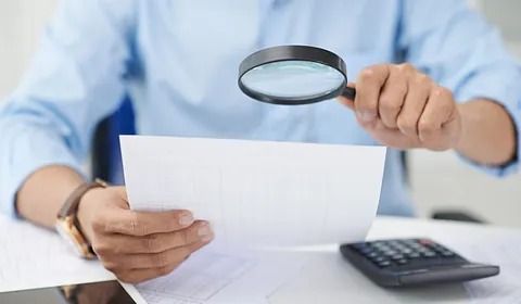 Una persona usa una lupa para examinar un documento. Hay una calculadora y un reloj visibles sobre el escritorio.