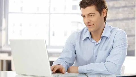 Hombre con camisa azul trabajando en una computadora portátil en un escritorio, sonriendo, oficina luminosa.