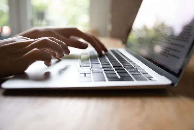 Manos escribiendo en el teclado de una computadora portátil, probablemente en el interior sobre una superficie de madera.