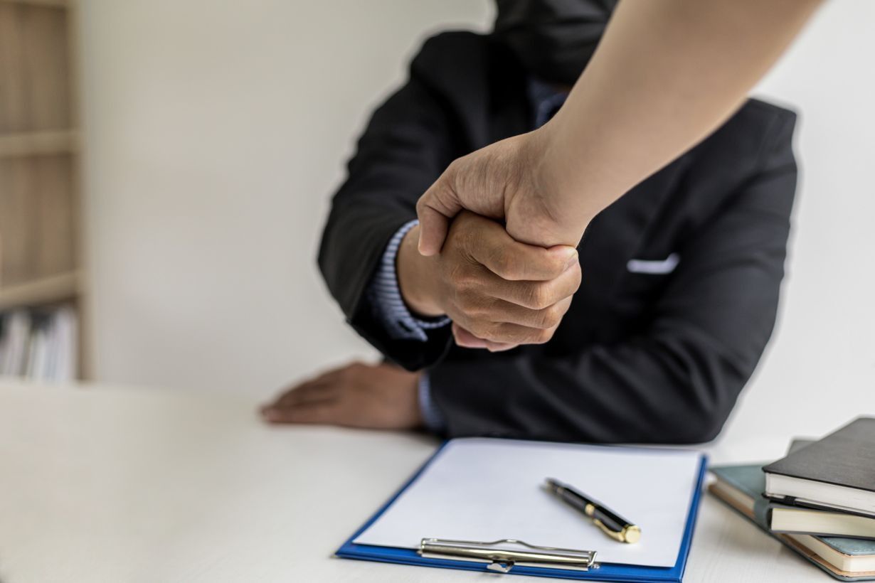 Apretón de manos entre dos personas vestidas de negocios sobre un documento y un bolígrafo en un escritorio.