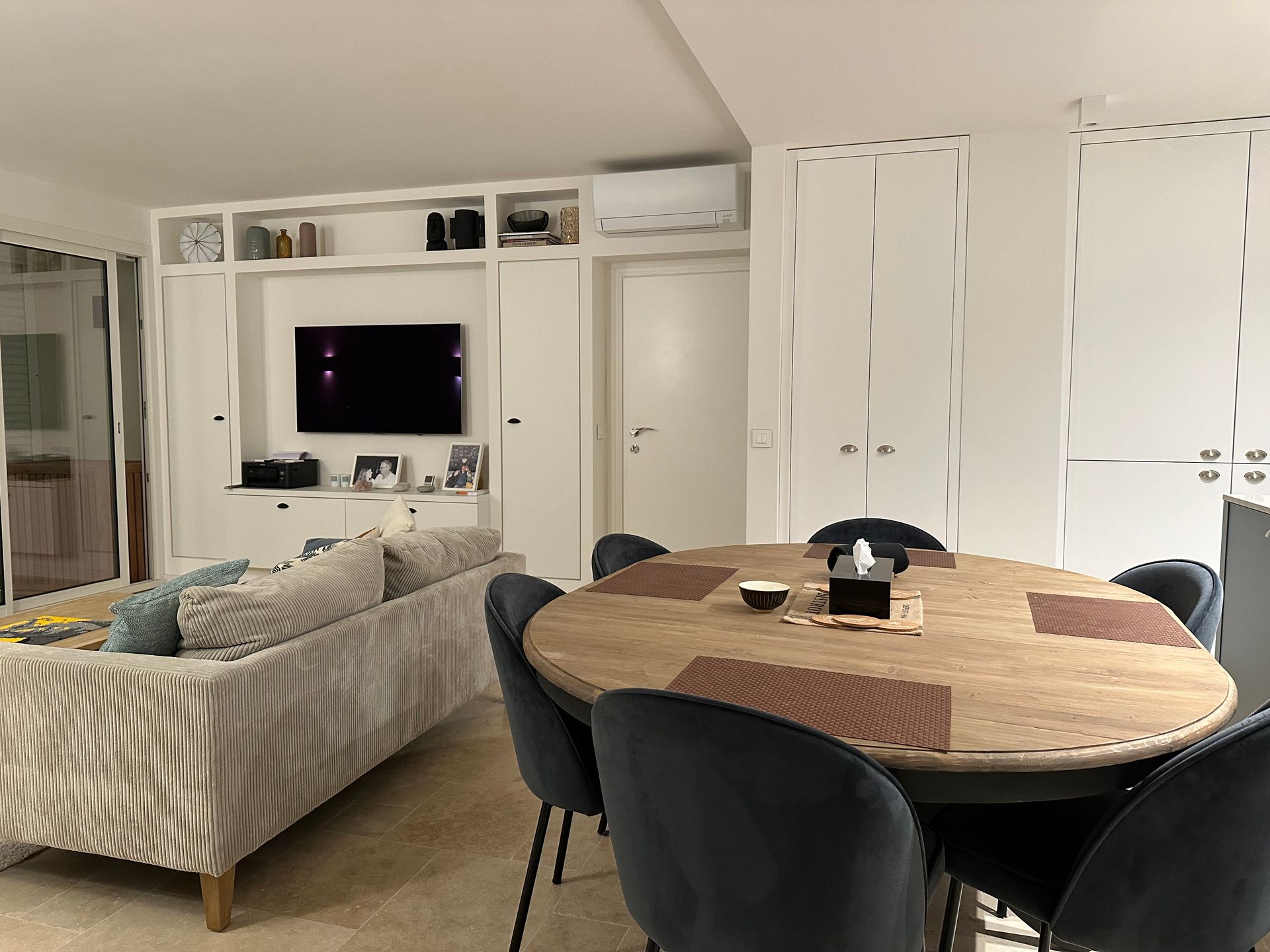Séjour avec canapé, table à manger ovale, placards intégrés et télévision. Chaises bleu foncé et table en bois.