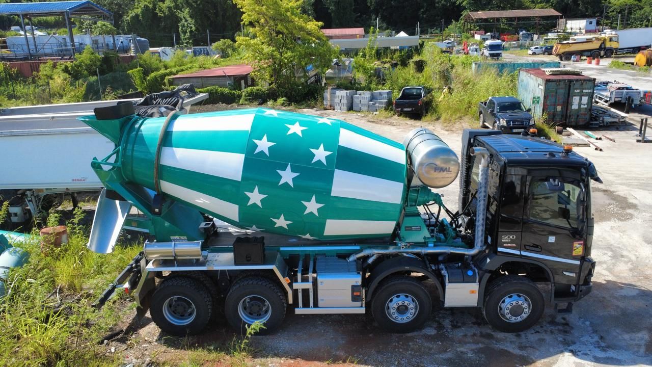 Un camion-malaxeur à béton déverse du ciment sur une allée de gravier ; deux ouvriers à proximité avec des outils.