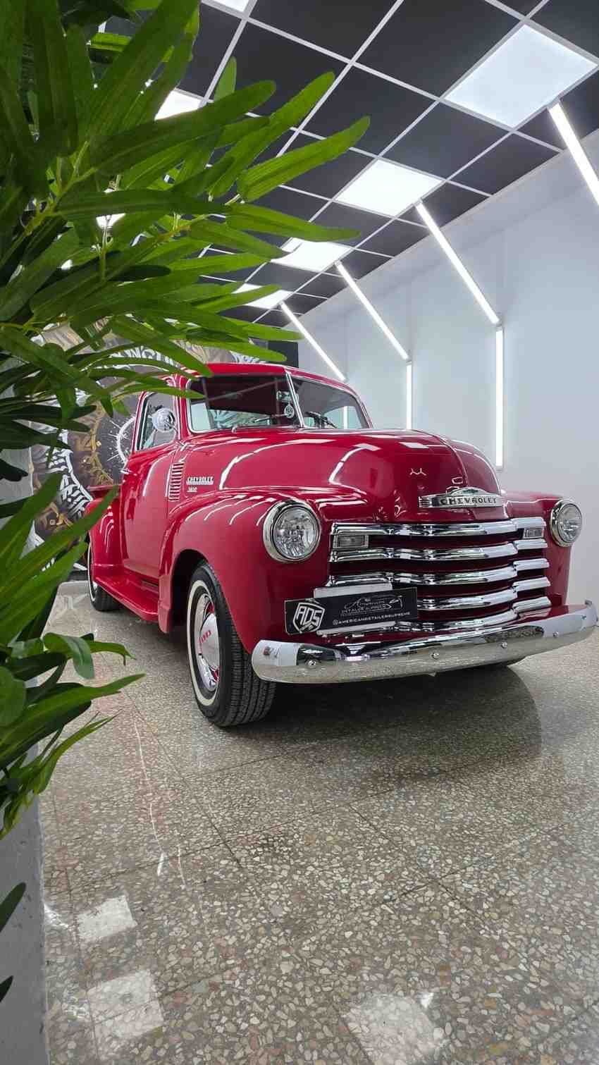 Un coche clásico rojo en la sala de exposición, enmarcado por plantas verdes y luces modernas en el techo.