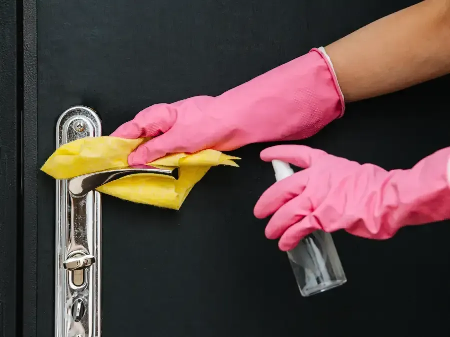 Una persona que lleva guantes rosas está limpiando la manija de una puerta con un paño y una botella de spray.