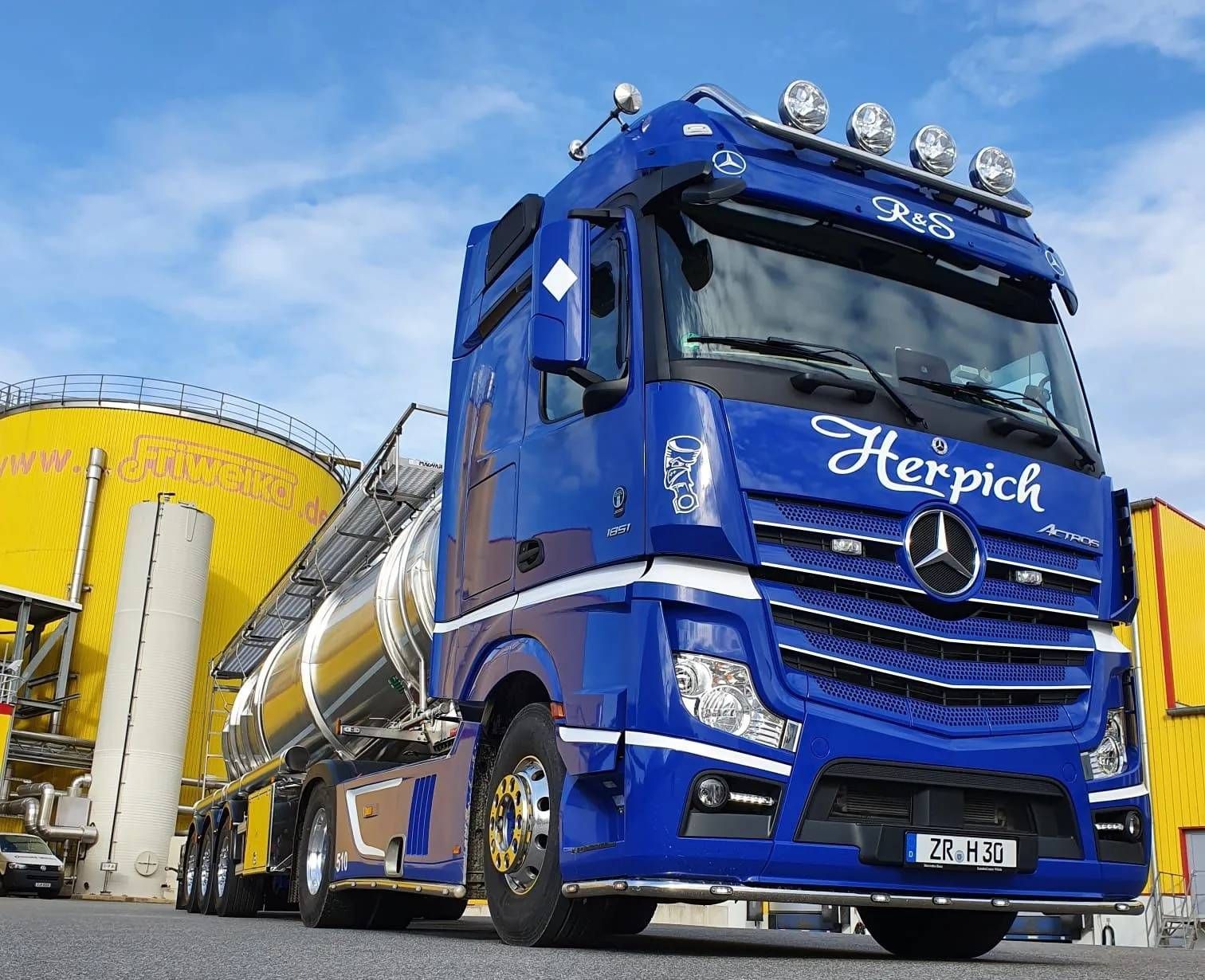 Blauer und silberner Tankwagen von Mercedes-Benz, der an einem sonnigen Tag in der Nähe eines gelben Industriesilos geparkt ist.