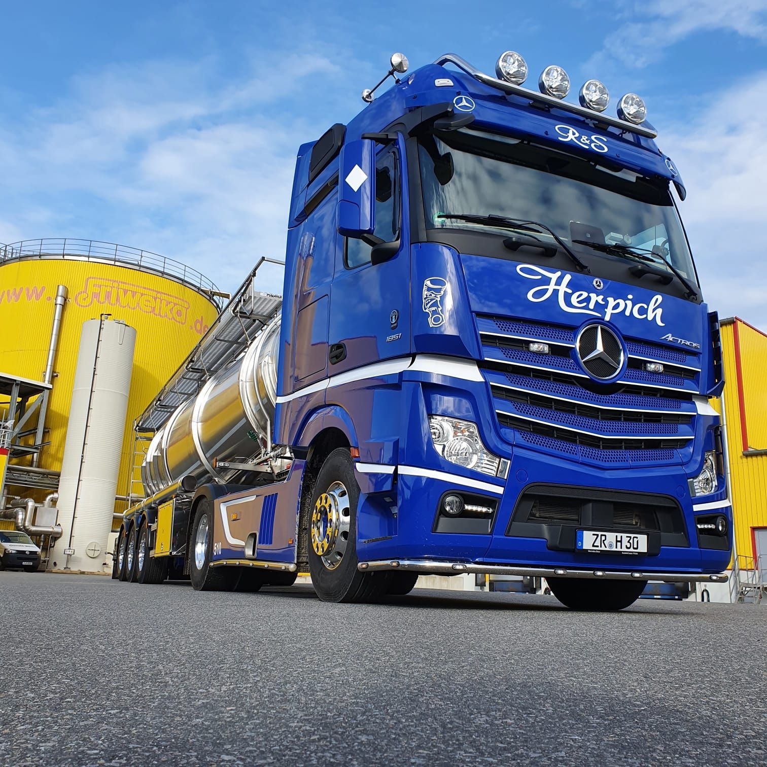 Blauer Mercedes-Sattelschlepper, der einen silbernen Tankwagen zieht; „Herpick“-Logo auf dem Führerhaus; geparkt in der Nähe eines gelben Gebäudes.