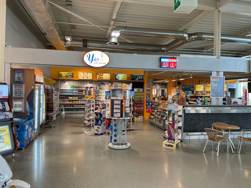 Intérieur d'une supérette avec rayons de snacks, caisse et sièges en métal près de l'entrée