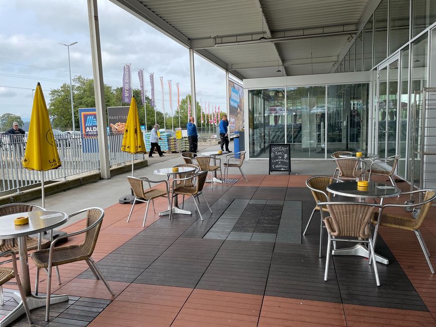 Terrasse de café avec tables et chaises en métal sous une allée couverte, à côté de parasols jaunes et de vitrines.