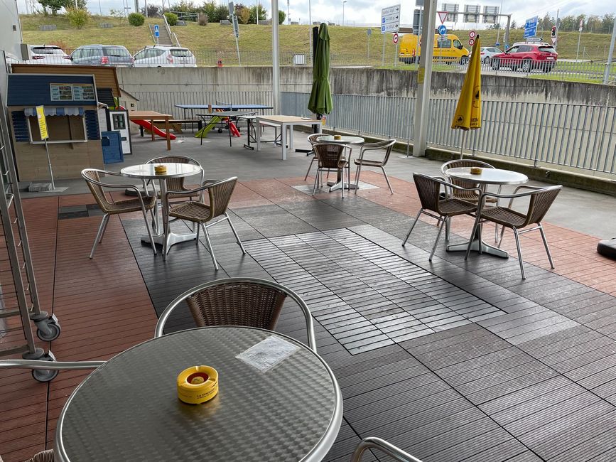 Terrasse de café en plein air avec tables et chaises en métal, terrasse en bois et parasols, en bordure de rue.
