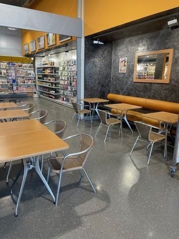 En arrière-plan, une terrasse de café vide avec des tables en bois, des chaises en métal et des étagères de snacks.