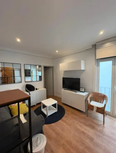 Salón moderno con televisor, armarios blancos, suelo de madera, estantería negra, silla y ventanas luminosas