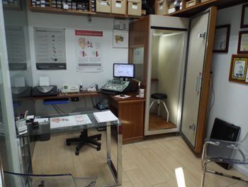 Pequeña oficina con escritorio, monitor de ordenador, silla, mapas en la pared y una puerta abierta a un pasillo estrecho.