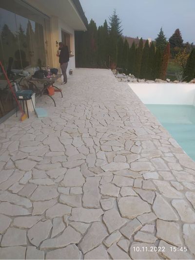 Steingepflasterte Terrasse neben einem türkisfarbenen Pool, mit Terrassenstühlen und hohen immergrünen Hecken am Rand.