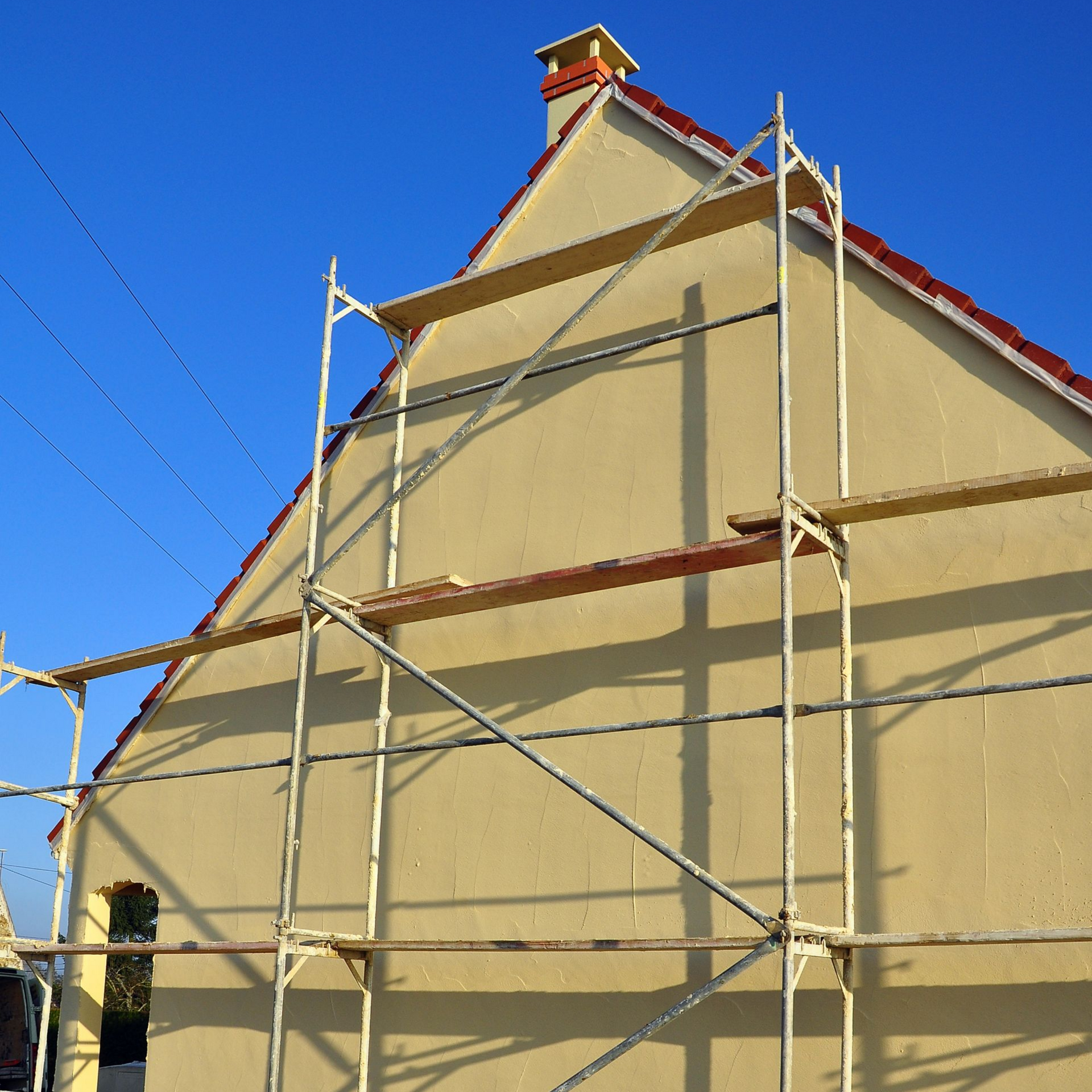 Échafaudages contre une maison aux murs beiges et au toit rouge sous un ciel bleu.