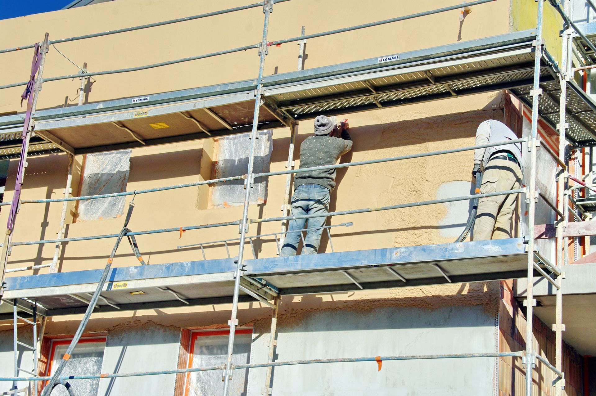 Deux ouvriers, juchés sur un échafaudage, appliquent du stuc sur un mur extérieur d'un bâtiment.