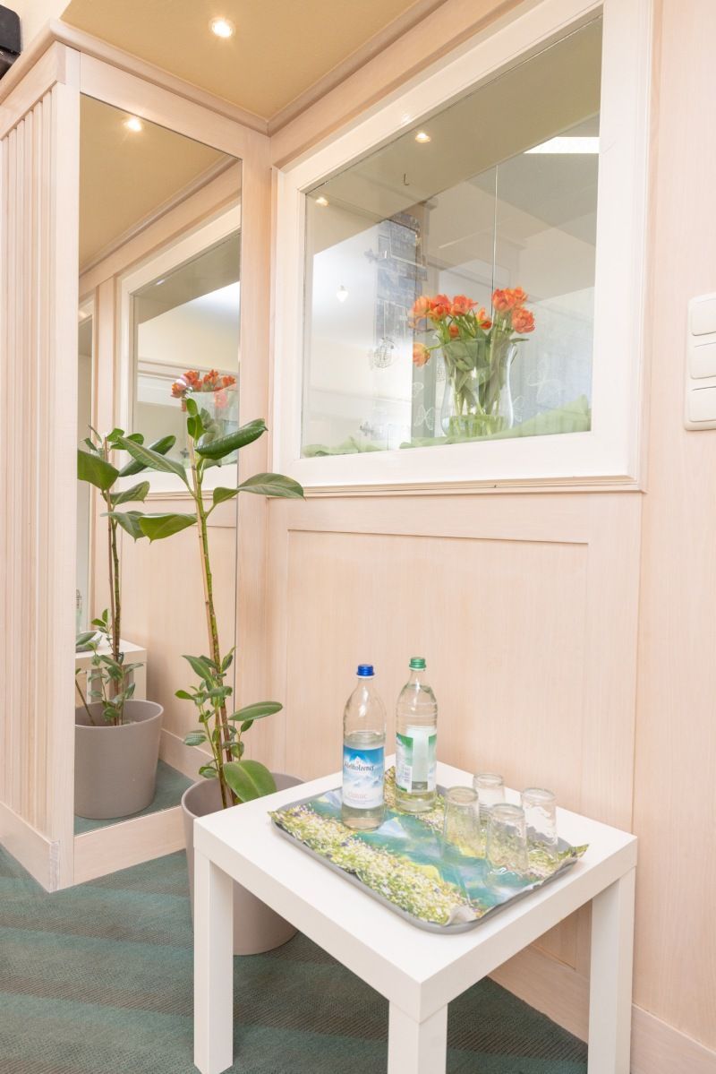 Ecke mit weißem Tisch, Wasserflaschen, Gläsern, Blumen in einer Vase, Spiegel und einer Pflanze. Rosa Wände, Fenster.