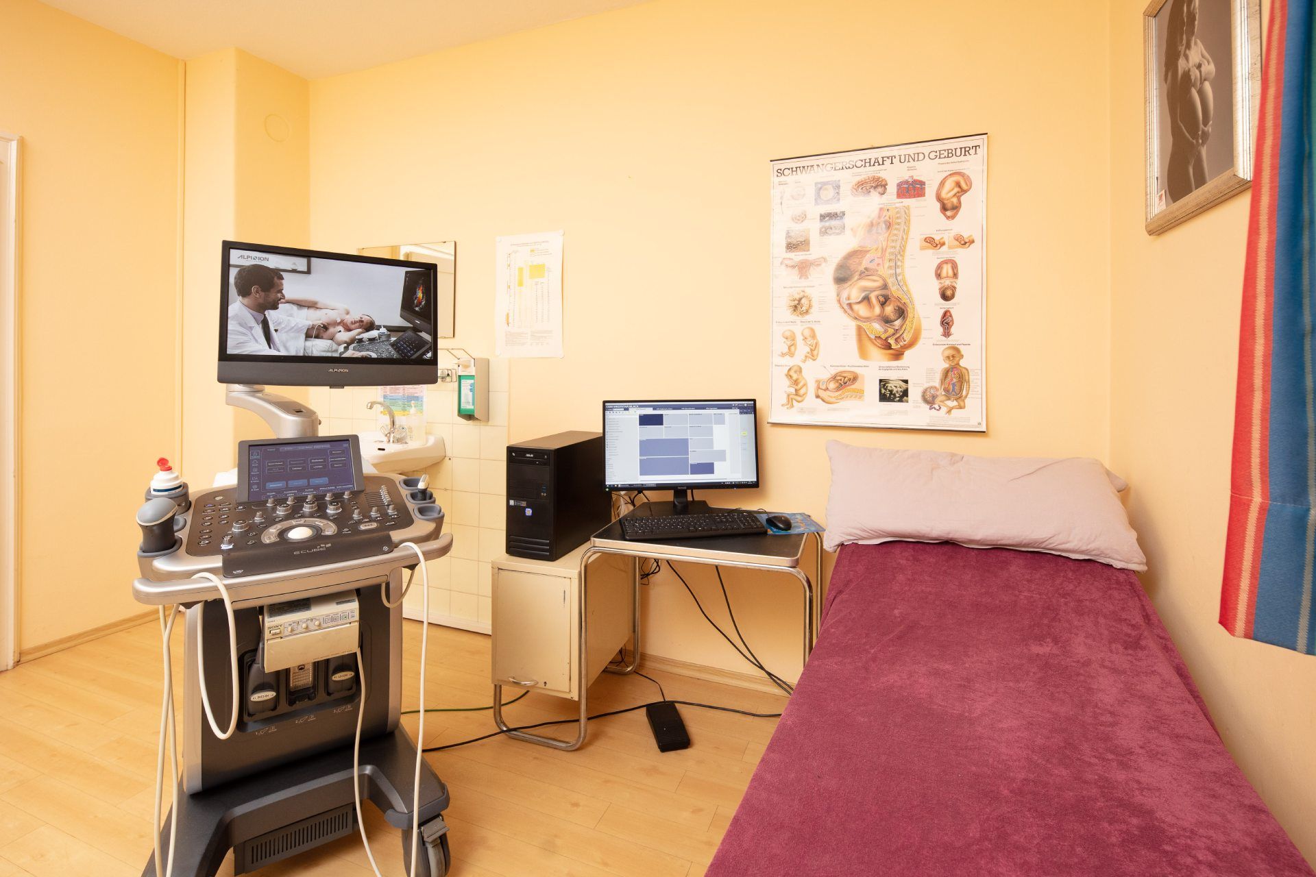 Untersuchungsraum: Ultraschallgerät mit Monitor, Computertisch, Untersuchungsliege, Anatomietafel an der Wand.