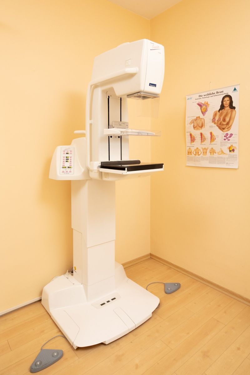 Ein Mammographiegerät in einem beigefarbenen Raum, neben einem Poster zum Thema Brustgesundheit.