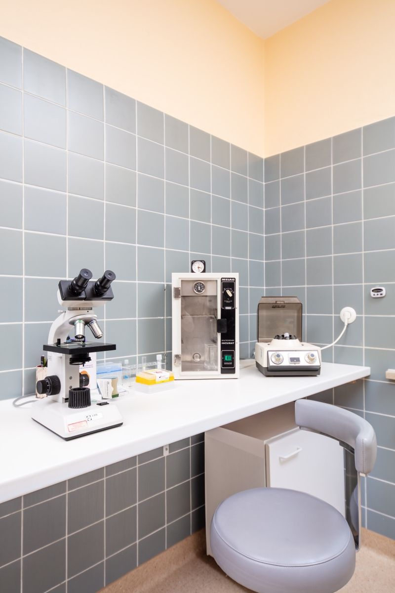 Labor mit Mikroskop, Inkubator, Zentrifuge und Hocker auf einer weißen Arbeitsfläche vor einer gefliesten Wand.