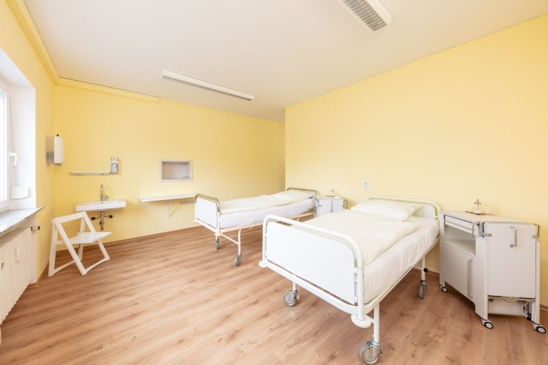 Krankenzimmer mit zwei Betten, einem Nachttisch, einem Waschbecken und einem Stuhl. Gelbe Wände und Holzboden.