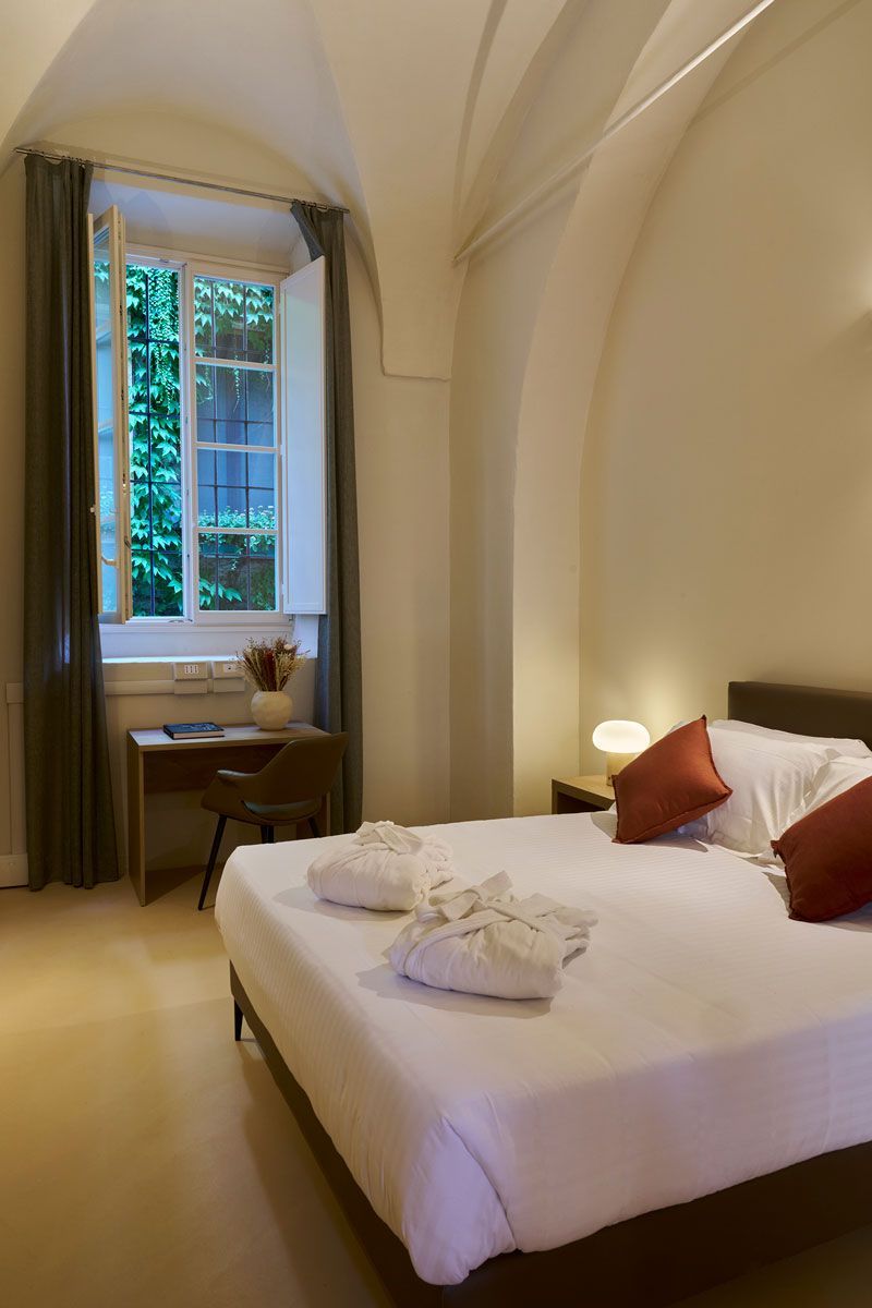 Camera matrimoniale con soffitto a volta, letto con cuscini bordeaux e accappatoi piegati, alta finestra affacciata sul verde e scrivania con fiori secchi.