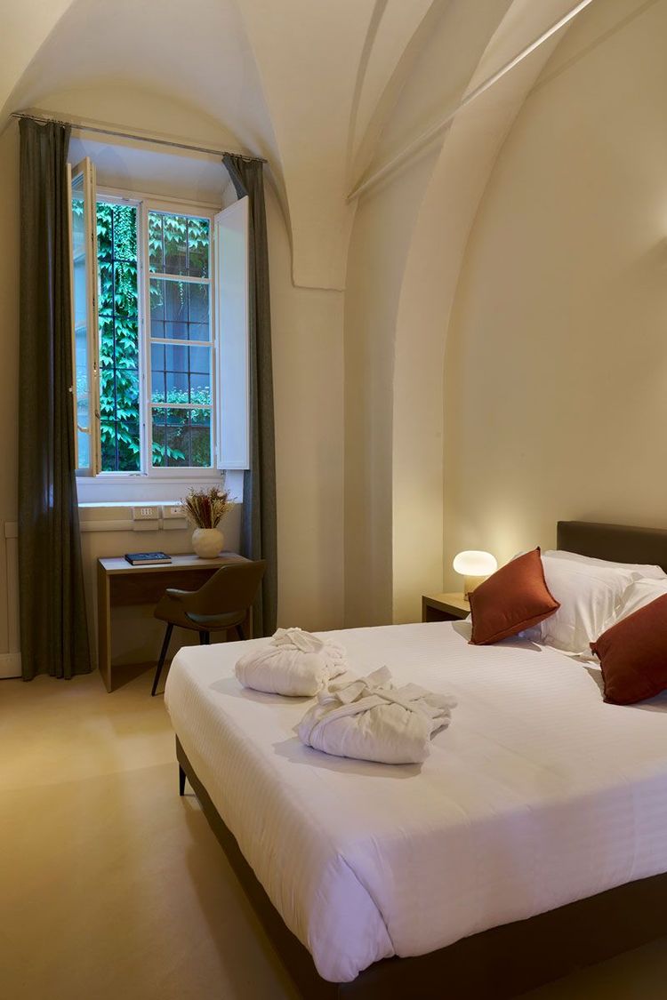 Camera matrimoniale con soffitto a volta, letto con cuscini bordeaux e accappatoi piegati, alta finestra affacciata sul verde e scrivania con fiori secchi.