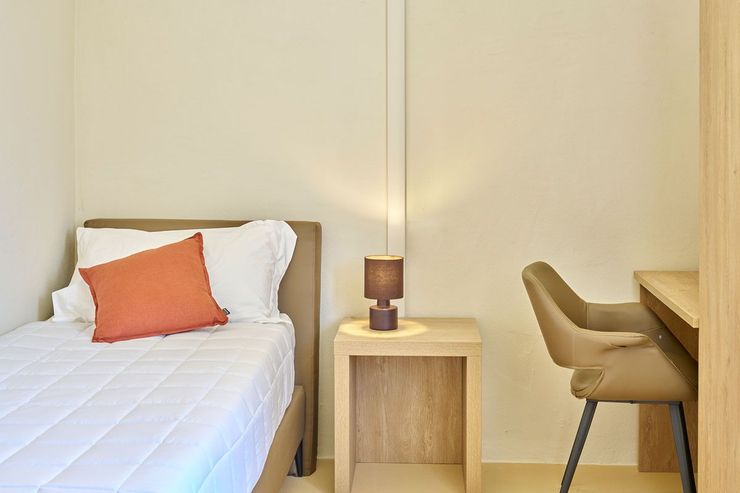 Camera singola con letto dal cuscino arancione, comodino in legno con lampada e scrivania con sedia.