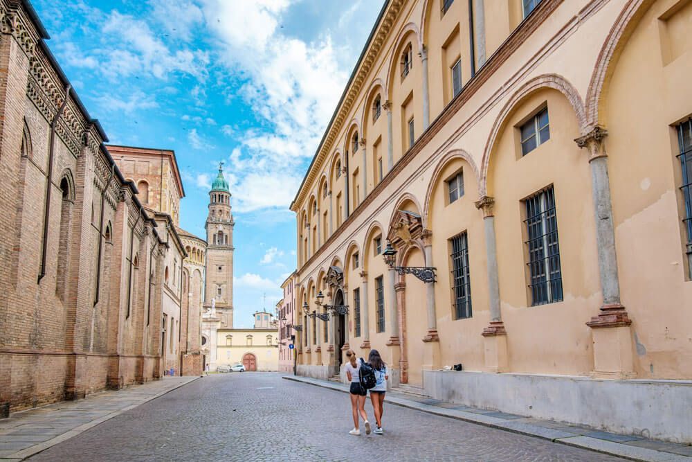 Scorcio del centro storico di Parma con un edificio porticato color ocra, un palazzo in mattoni e un campanile dalla cupola verde sotto un cielo azzurro.