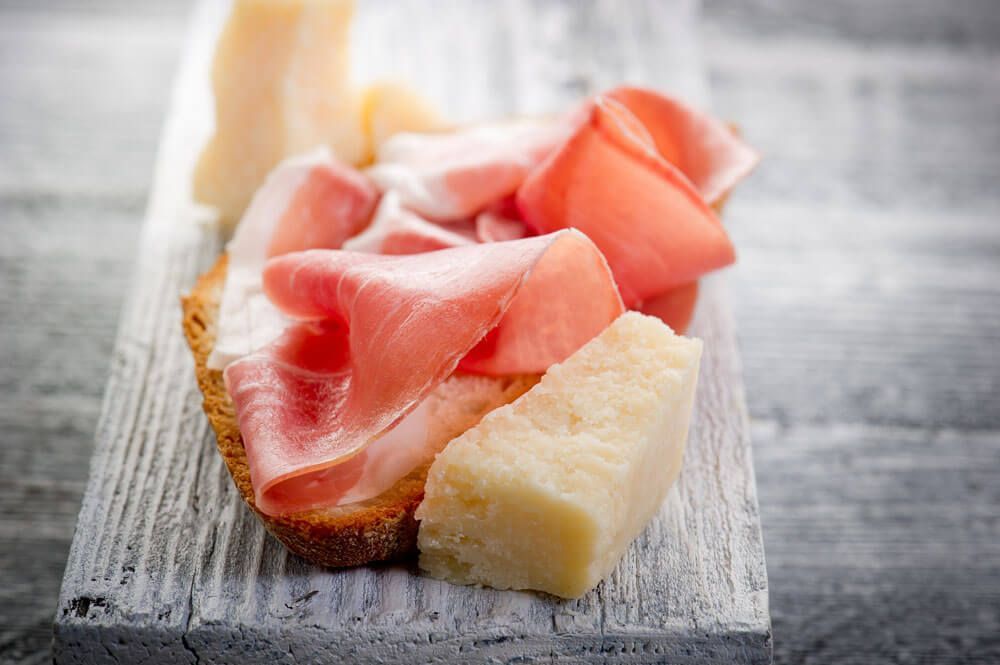 Fetta di pane con Prosciutto di Parma e una scaglia di Parmigiano Reggiano su un tagliere di legno.