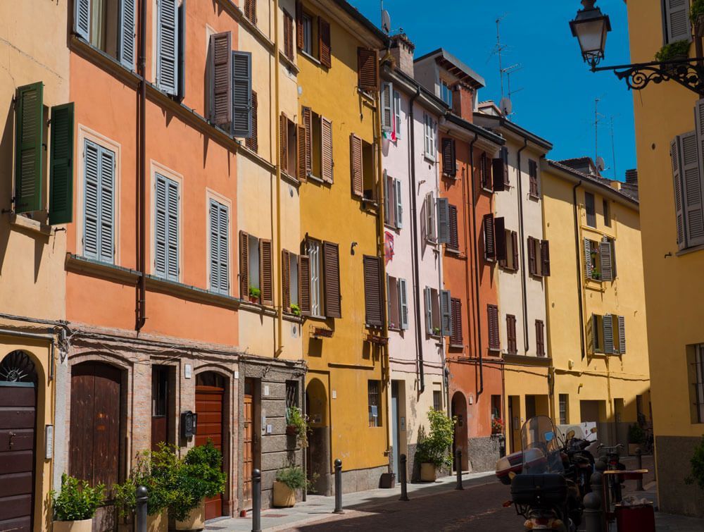 Borgo del centro storico di Parma con case dalle facciate colorate in arancio, giallo e rosa, persiane in legno e lampione in ferro battuto sotto un cielo azzurro.