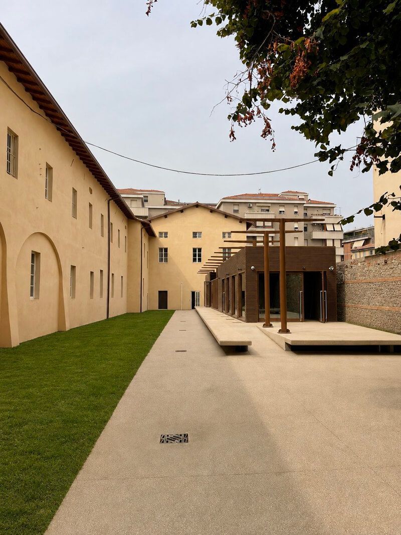 Facciata esterna di Chiostro 26 color ocra, con finestre ad arco, prato verde, vialetto e pergolato in legno.