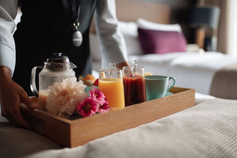 Un membro dello staff porta in camera un vassoio in legno con la colazione: spremuta d'arancia, succo rosso, teiera, tazza e fiori freschi, appoggiato sul letto.